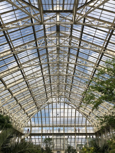 Interior view of a spacious, well-maintained shopping arcade with a high, curved glass ceiling supported by ornate white columns with gold and red accents. The tiled floor features a decorative blue a